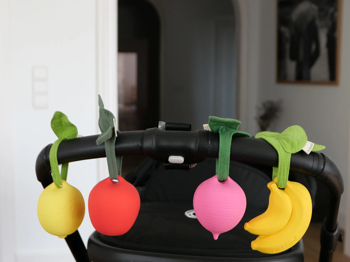 Oli and Carol fruit and vegetable stroller toys hanging from a black handlebar