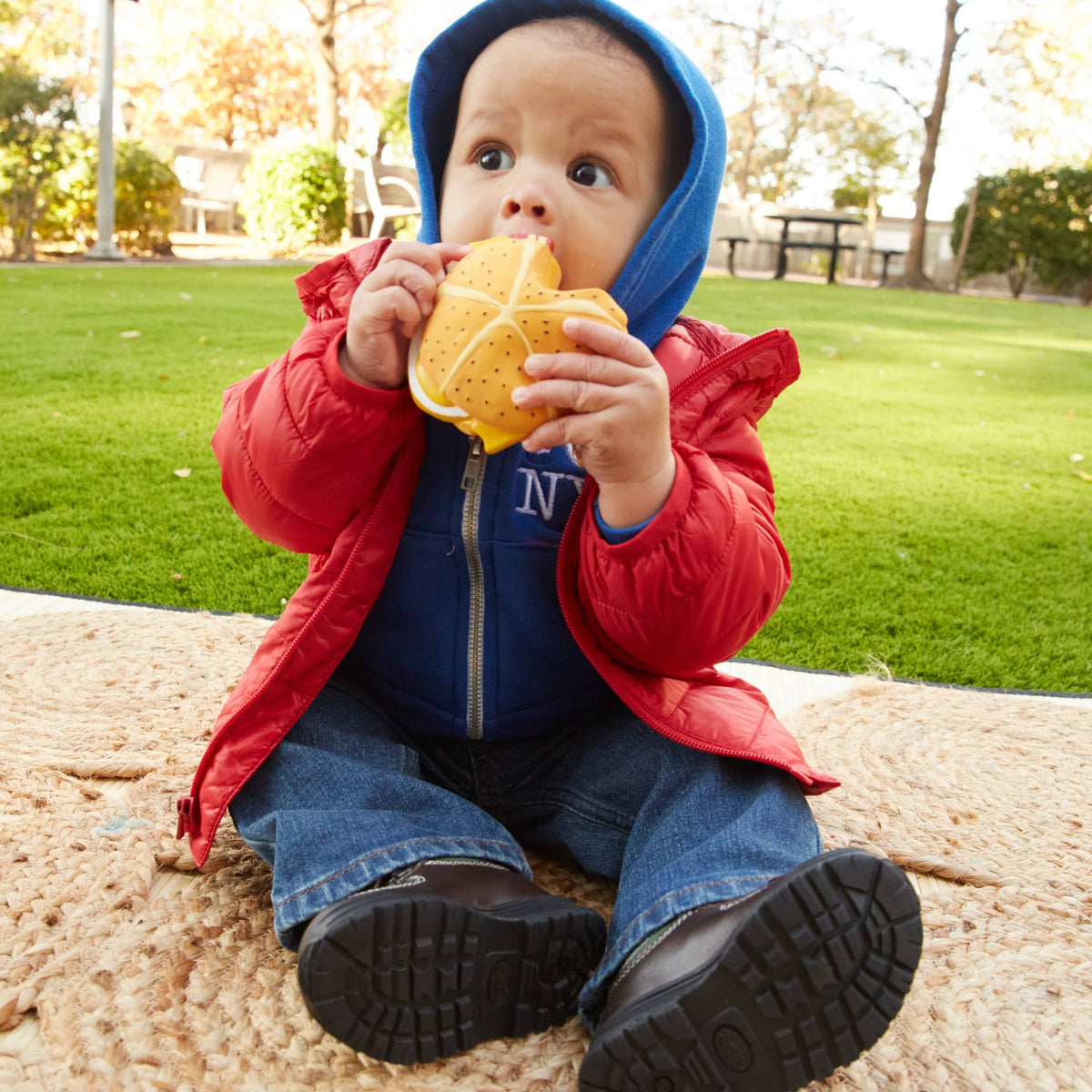 Toddler in a red jacket and blue hoodie sitting on a mat chewing on a yellow toy burger