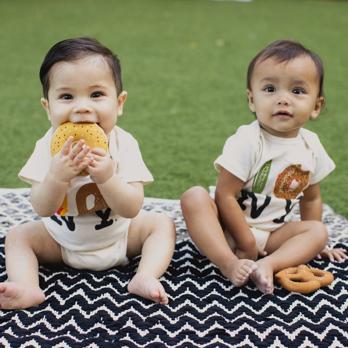Two babies sitting on a patterned blanket with bagel shaped teethers