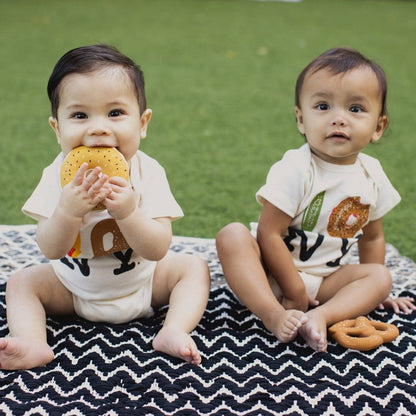 Two babies sitting on a patterned blanket with bagel shaped teethers