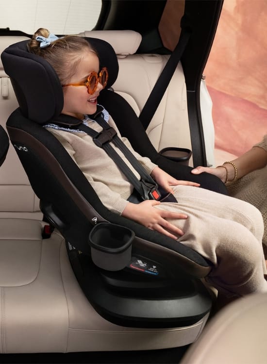 Girl wearing sunglasses smiles in black car seat with cup holders, beige car interior, adult hand nearby.