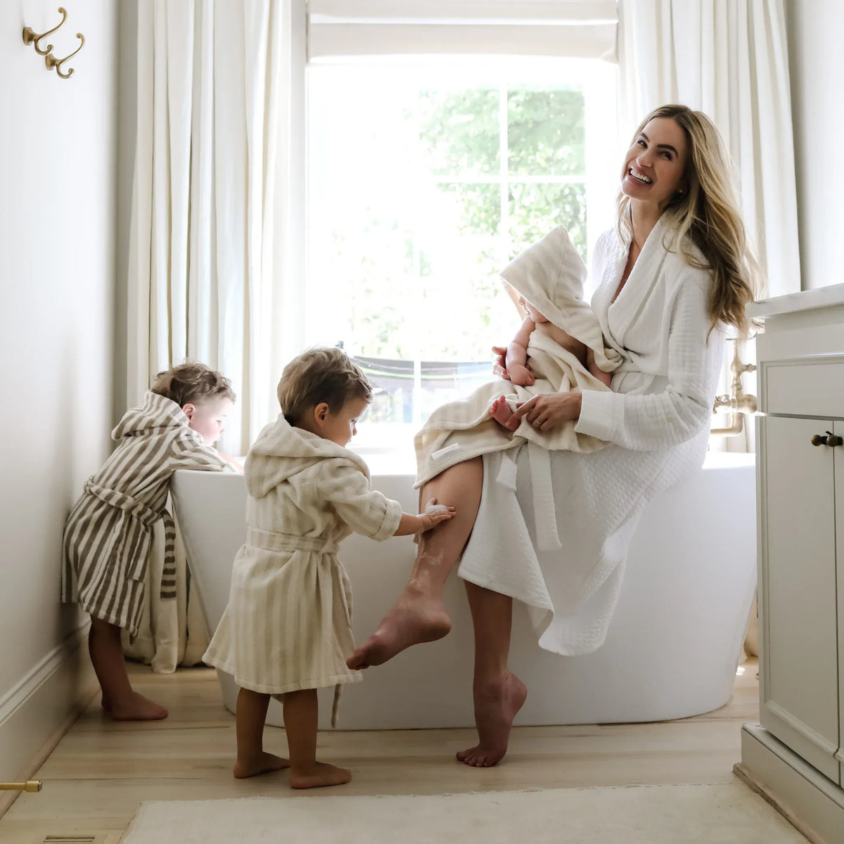 Woman in white robe sitting on bathtub with baby and two toddlers in striped robes