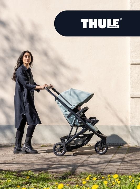 Woman in dark coat pushes light blue Thule stroller on sunny sidewalk, yellow flowers in foreground, brand logo top right.