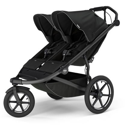 Black double jogging stroller by Thule with magnetic buckle, side view, large all-terrain wheels, padded seats, and white trim accents