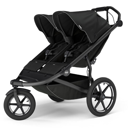 Black double jogging stroller by Thule with magnetic buckle, side view, large all-terrain wheels, padded seats, and white trim accents