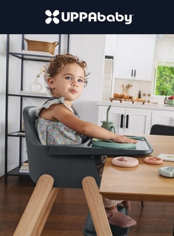 Toddler in gray UPPAbaby high chair at wooden table, wearing floral dress, reaching for green plate, kitchen background.