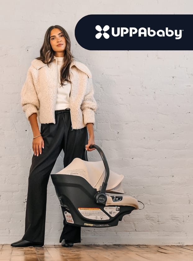 Woman in beige jacket and black pants stands beside UPPAbaby infant car seat, holding handle, against white brick wall.