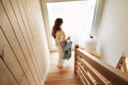 Woman carrying a folded sage green baby high chair on a wooden staircase