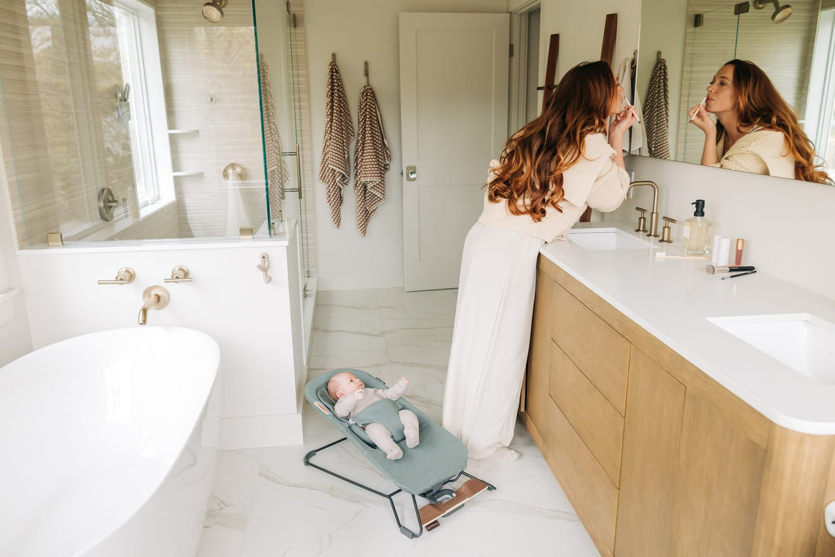 Woman applying makeup at bathroom vanity while baby sits in a bouncer