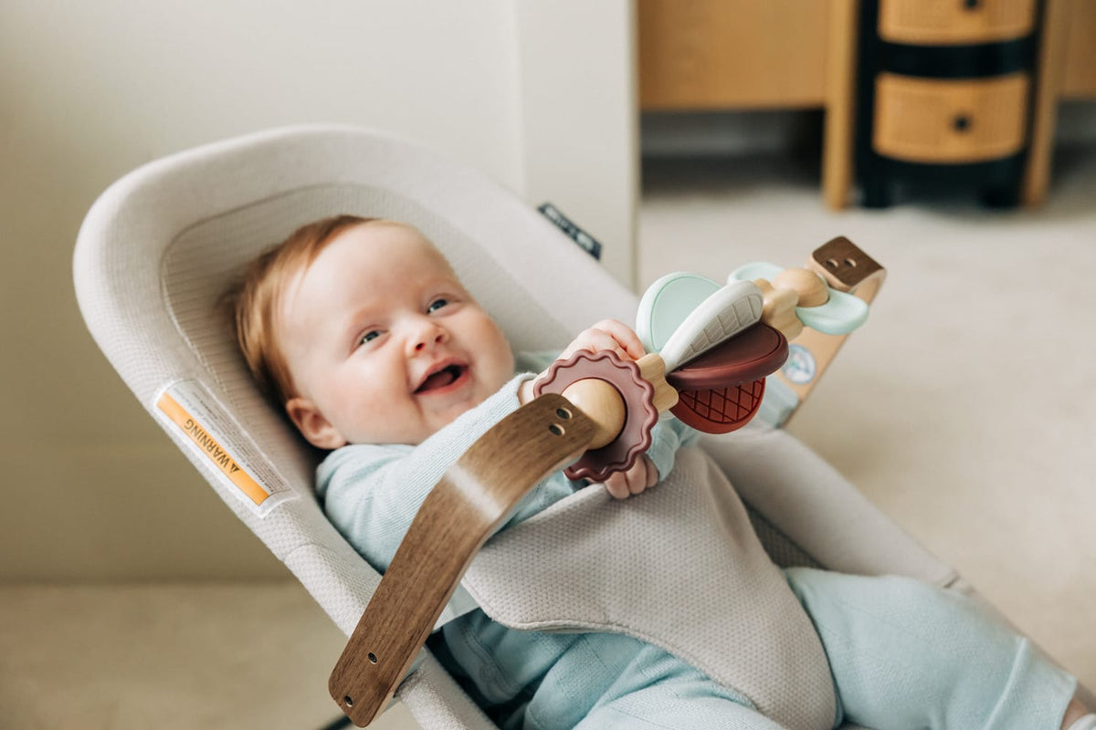 Baby sitting in a beige bouncer seat with a wooden toy bar attached