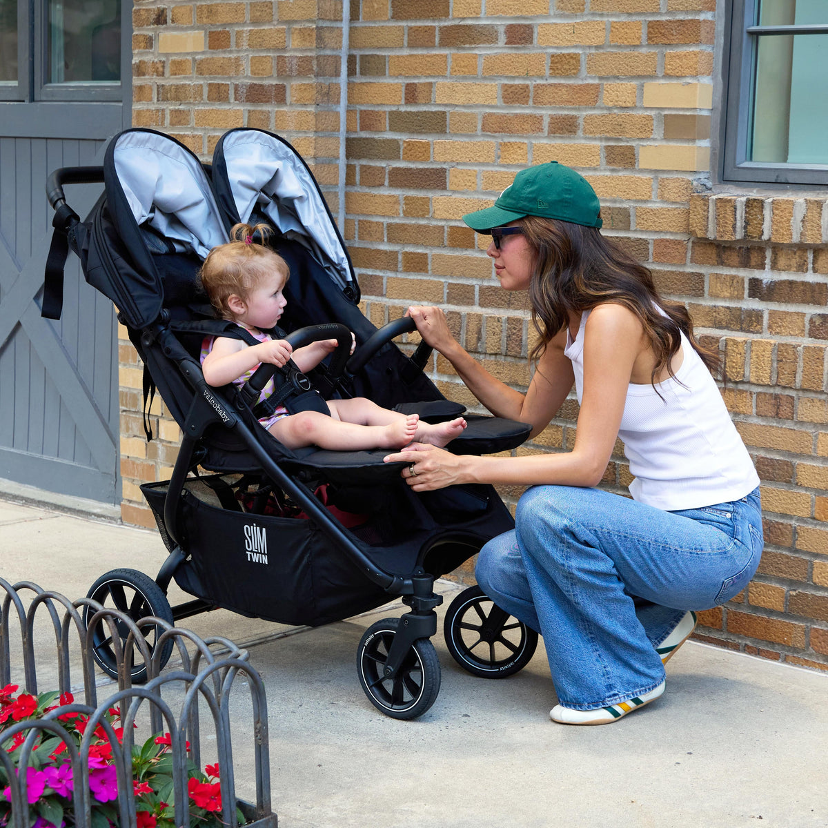 Woman kneeling beside a black Valco Slim Twin double stroller with a toddler seated inside